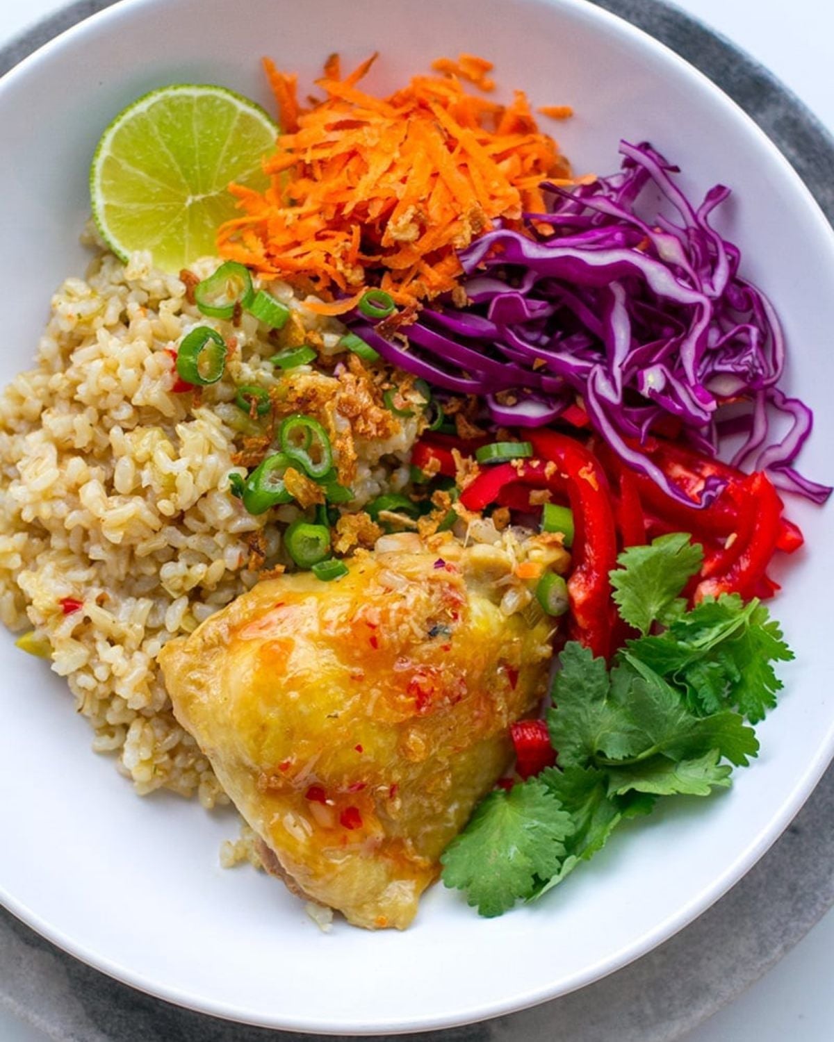 An overhead view of a white bowl filled with cooked brown rice and a tender piece of chicken, accompanied by shredded carrots, purple cabbage, sliced red bell peppers, fresh cilantro, chopped green onions, and a lime wedge.
