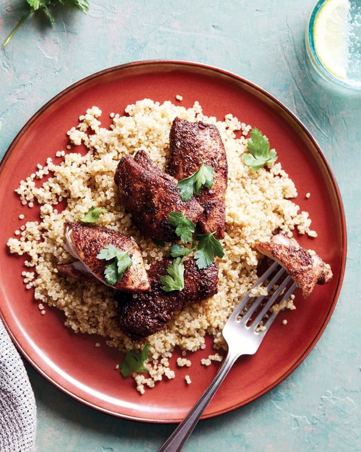 An overhead view of a red plate filled with fluffy cooked quinoa, topped with pieces of spiced, browned jerk chicken and garnished with fresh green parsley leaves. A silver fork rests on the plate next to a piece of chicken, and a glass with a lemon slice is visible in the upper right corner.