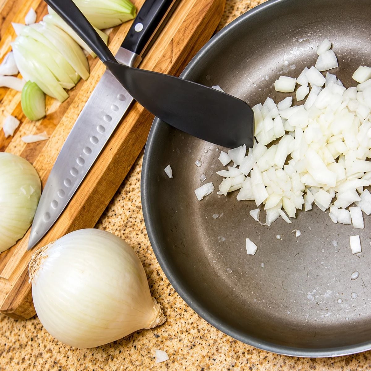 Chopped onion sits in a skillet on a kitchen counter beside a whole onion and chopping board