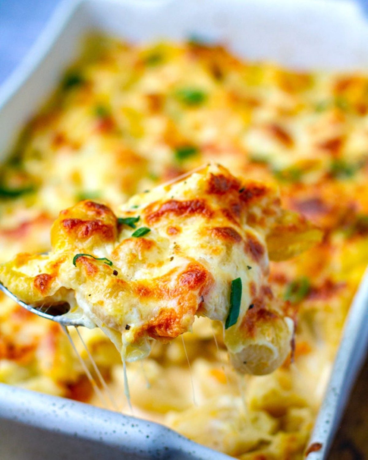 A spoonful of cheesy pumpkin pasta bake being lifted from a baking dish, highlighting a golden-brown, melted cheese crust and herbs.