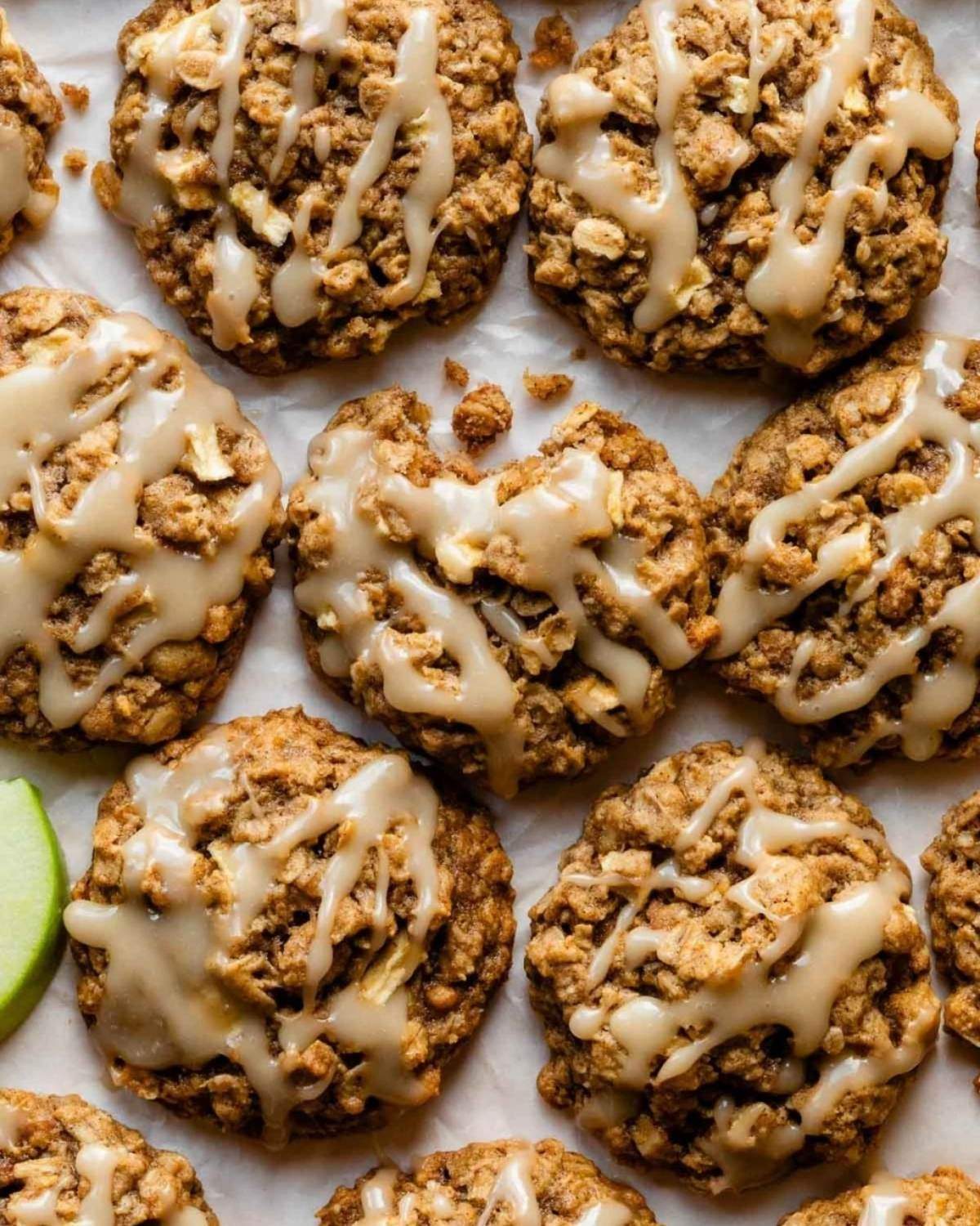 A flat lay of freshly baked apple cinnamon oatmeal cookies drizzled with a sweet white glaze, arranged on parchment paper with a piece of green apple visible on the side.