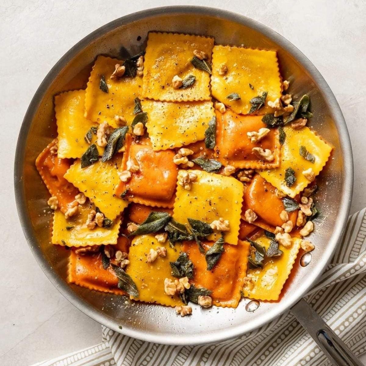 Square stuffed pasta pockets tossed with crunchy walnut pieces and crispy sage leaves inside a metal pan.
