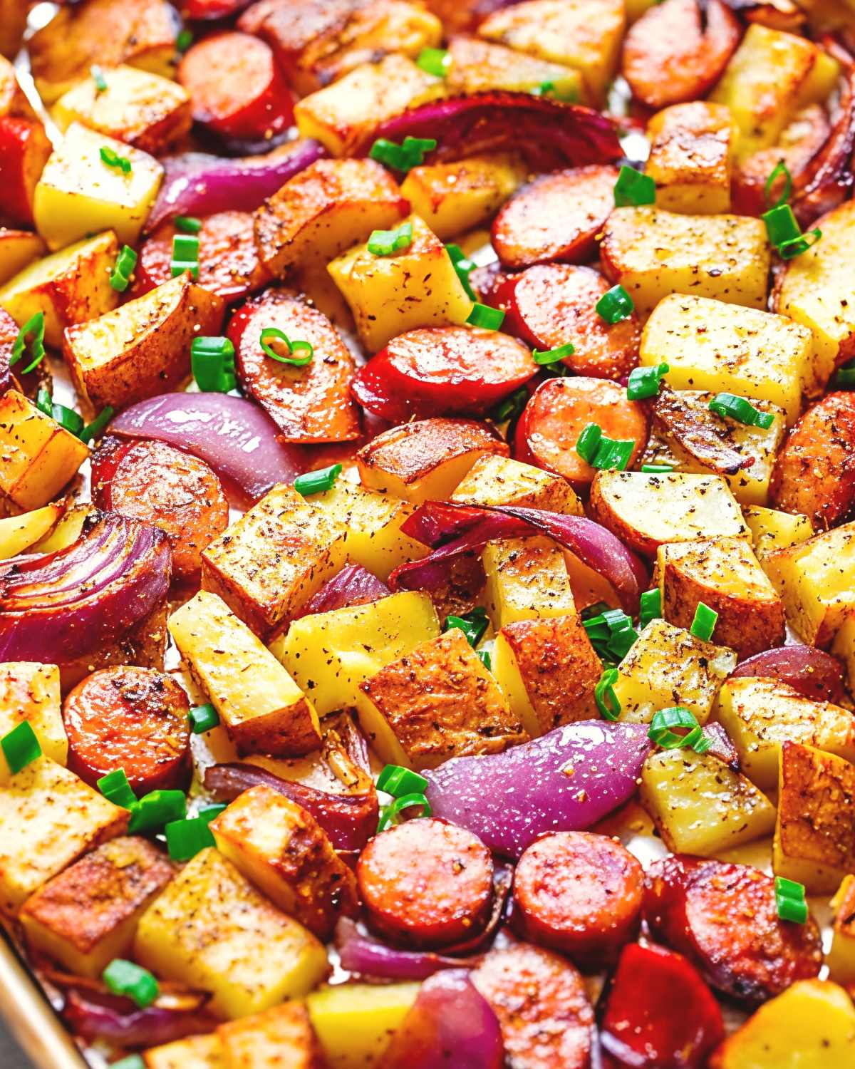 A close-up shot of roasted sausage, potatoes, and red onions on a pan, garnished with chopped green onions for an easy fall dinner.