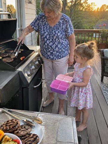 At the back porch grill just before dinner, a woman turns food with tongs while a small grandchild stands beside her holding an open lunchbox, neither looking at the camera.