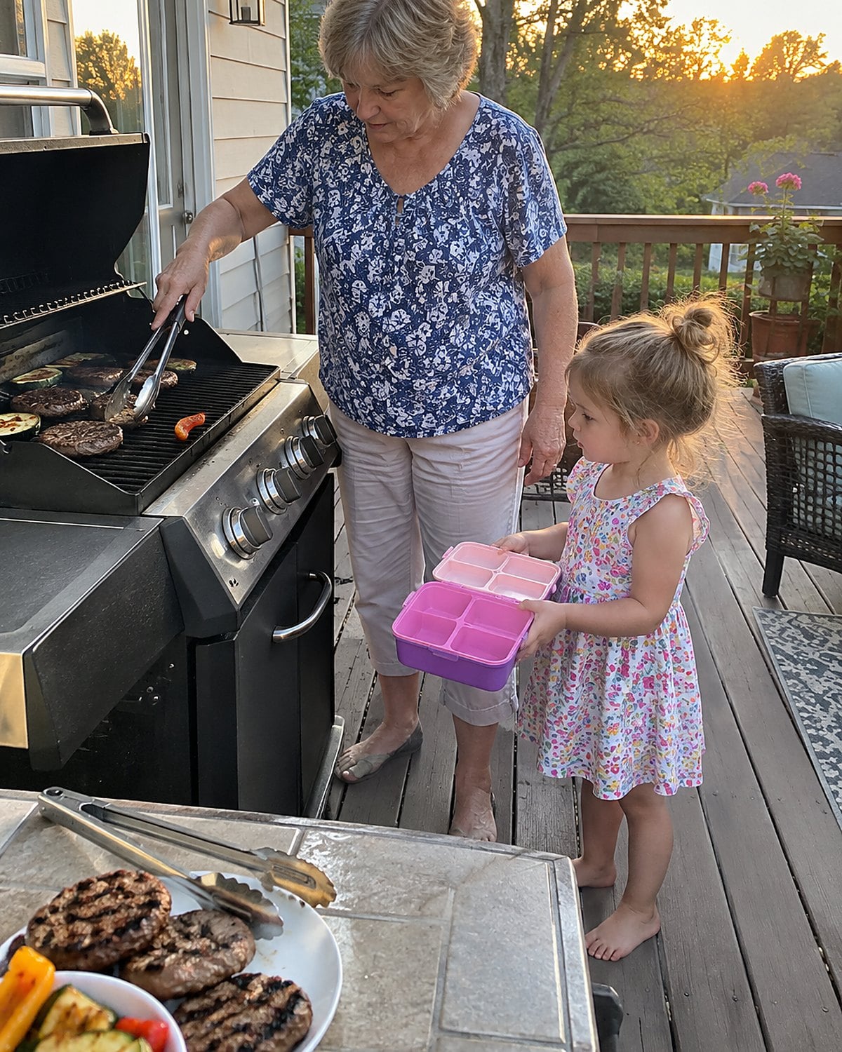 At the back porch grill just before dinner, a woman turns food with tongs while a small grandchild stands beside her holding an open lunchbox, neither looking at the camera.