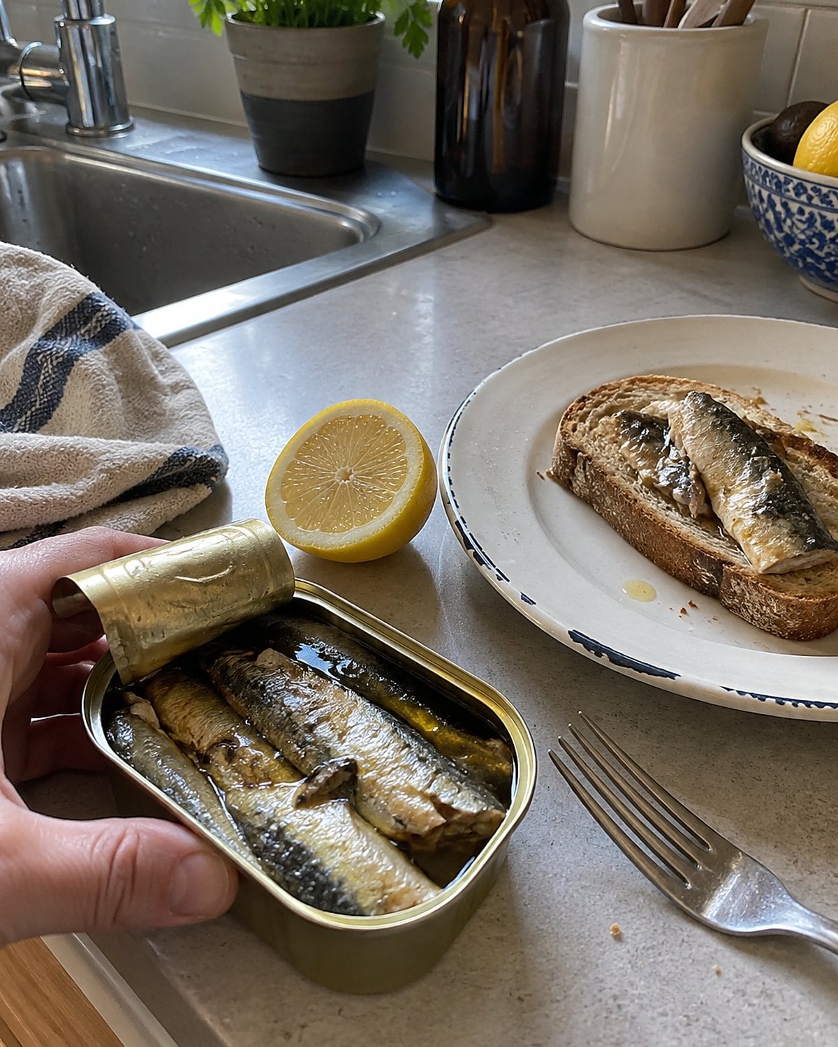 An opened sardine tin, a lemon half, and a fork beside toast at the kitchen sink.