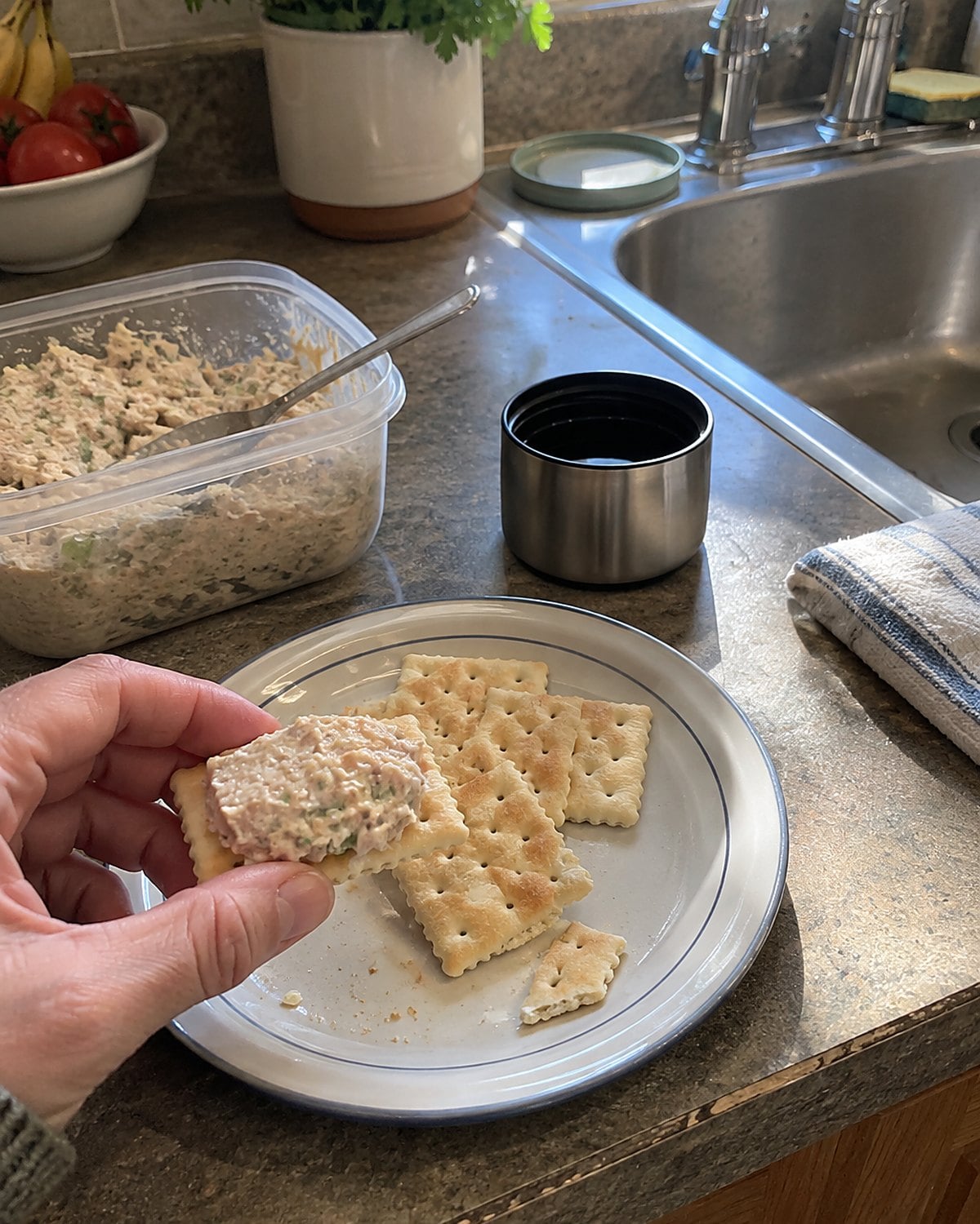 A small thermos cap and a sealed container of tuna salad beside crackers at the sink.