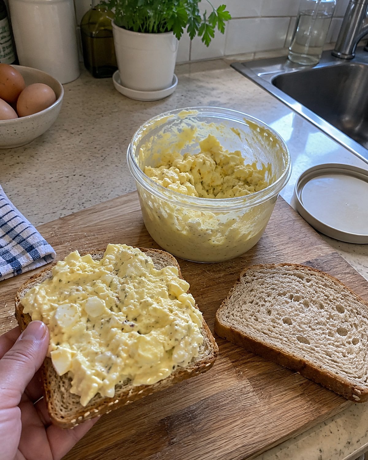 Egg salad in a lidded container beside separate slices of bread on a worn cutting board.
