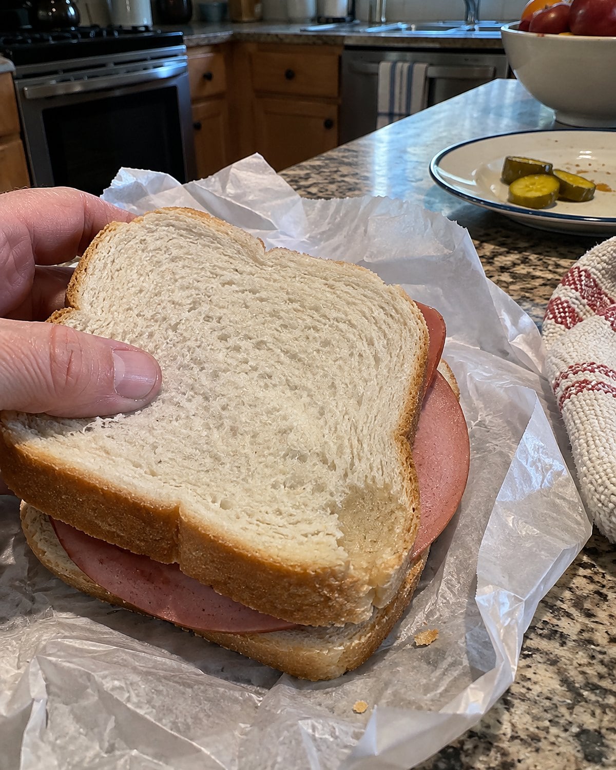 A bologna sandwich on soft white bread partly unwrapped on a kitchen counter, with a damp mark on the bread.