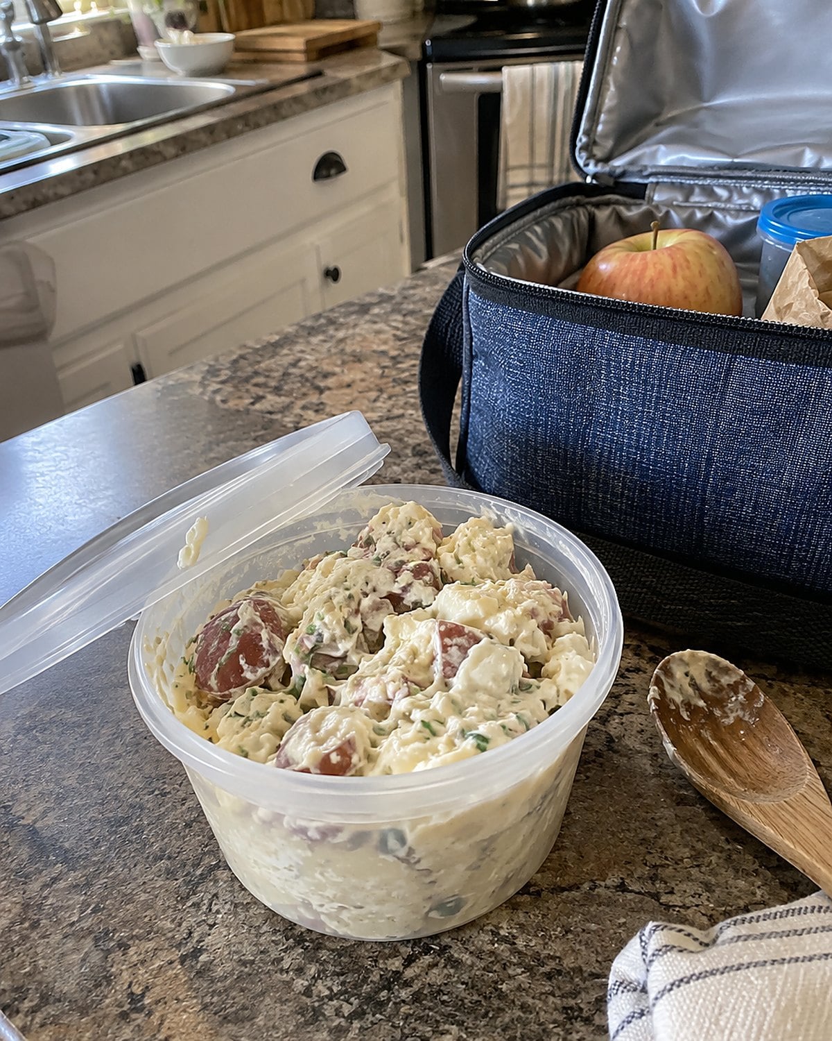 Potato salad in a small lidded cup beside an open insulated lunch bag on the counter.