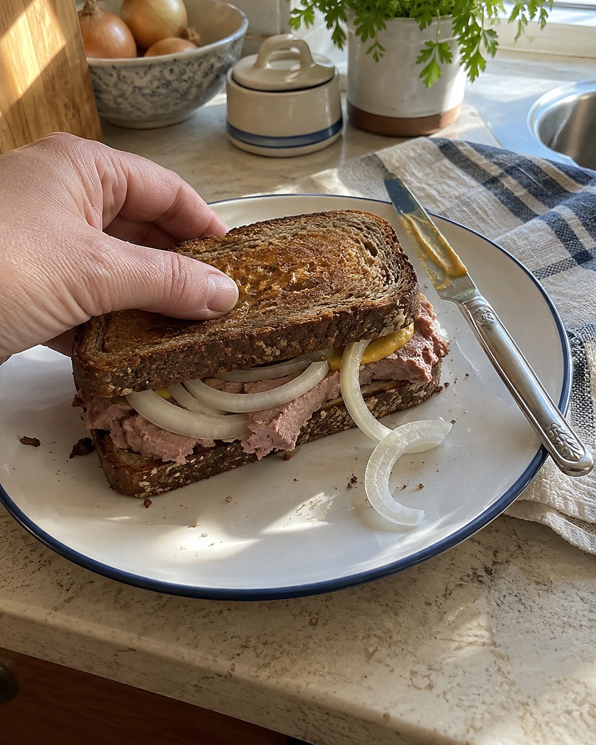 A liverwurst and onion sandwich on toasted rye on a kitchen counter, with mustard on the knife.