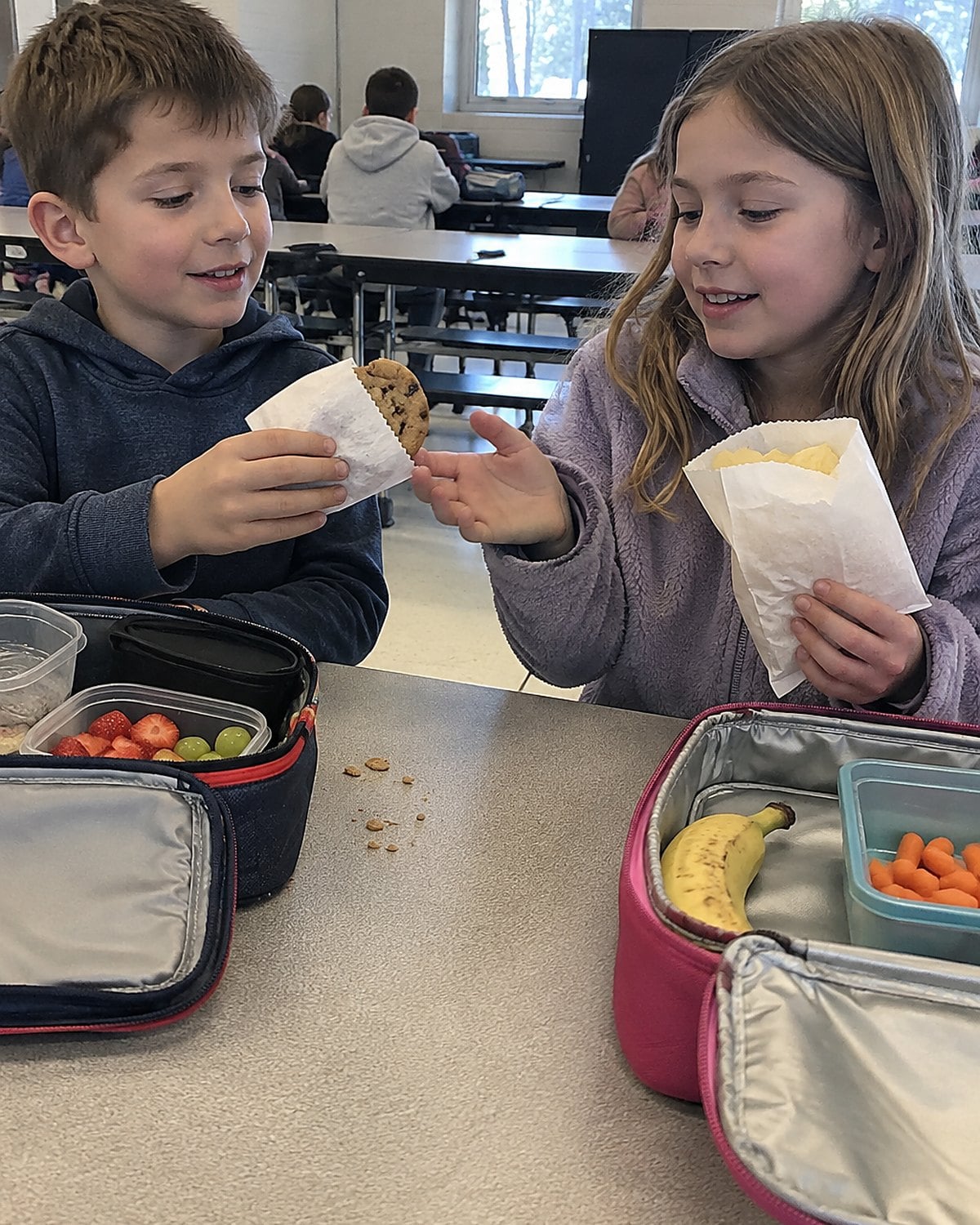 Two children's hands exchanging a wrapped cookie and a bag of chips above a school lunch table.
