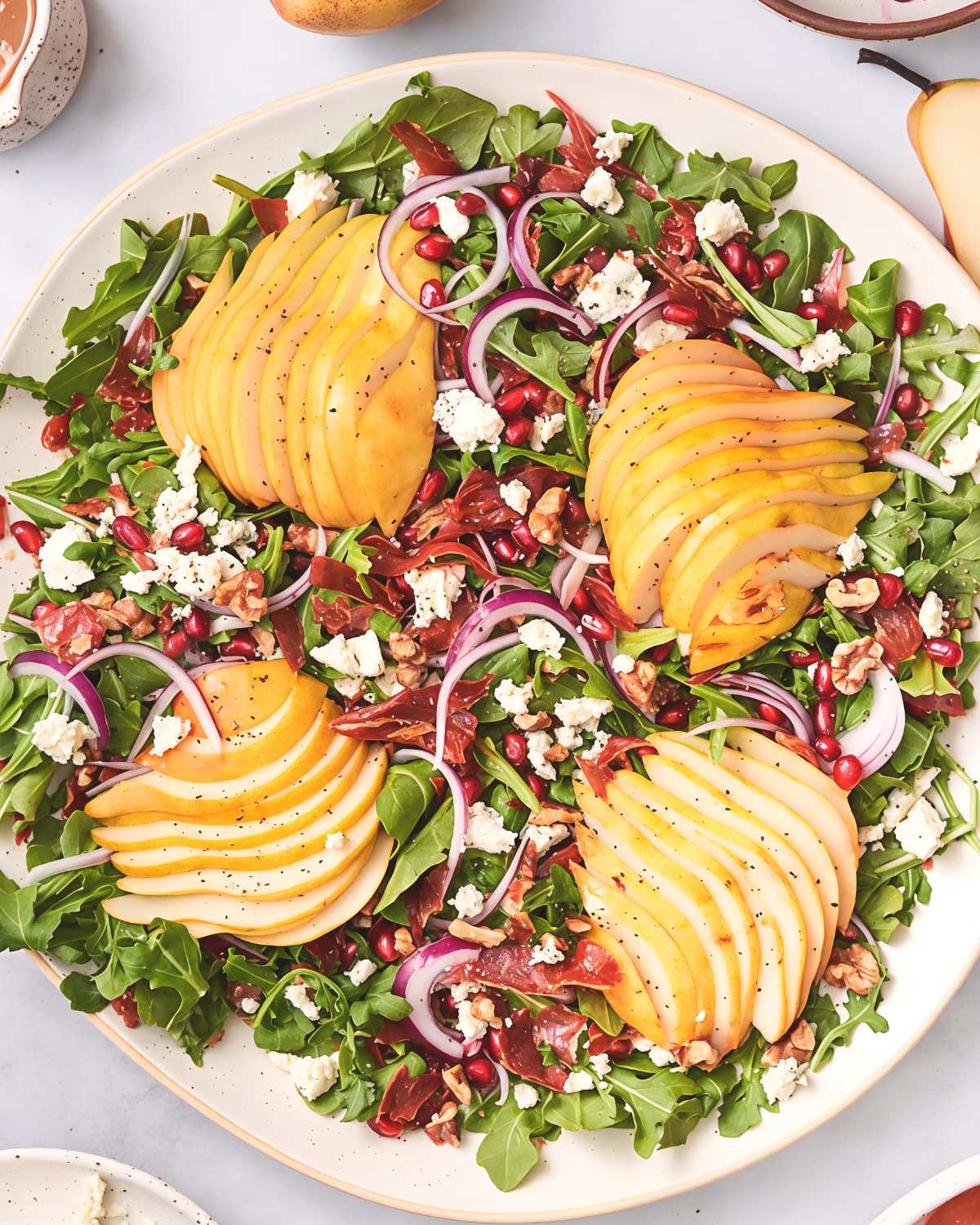 A colorful platter of arugula salad topped with sliced pears, pomegranate seeds, red onion rings, crumbled goat cheese, and chopped walnuts.