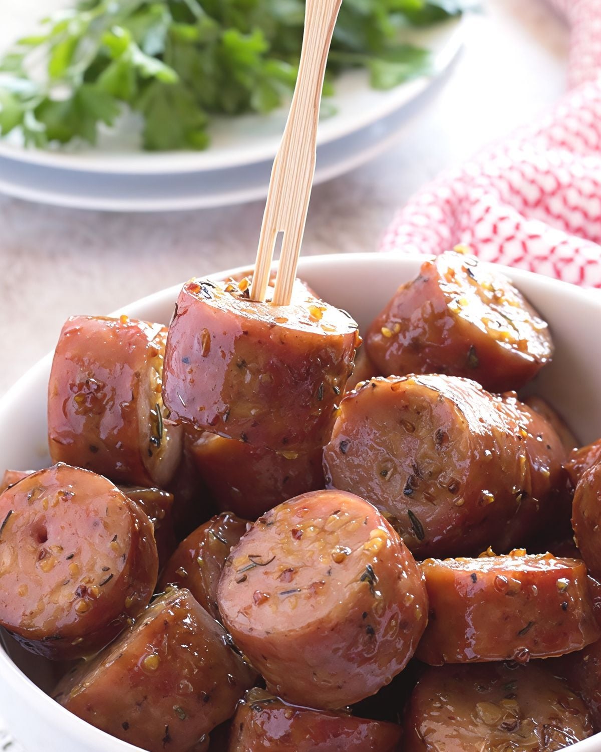 A wooden pick lifts a bite-sized slice of chicken apple sausage coated in a glossy maple mustard glaze from a white serving bowl