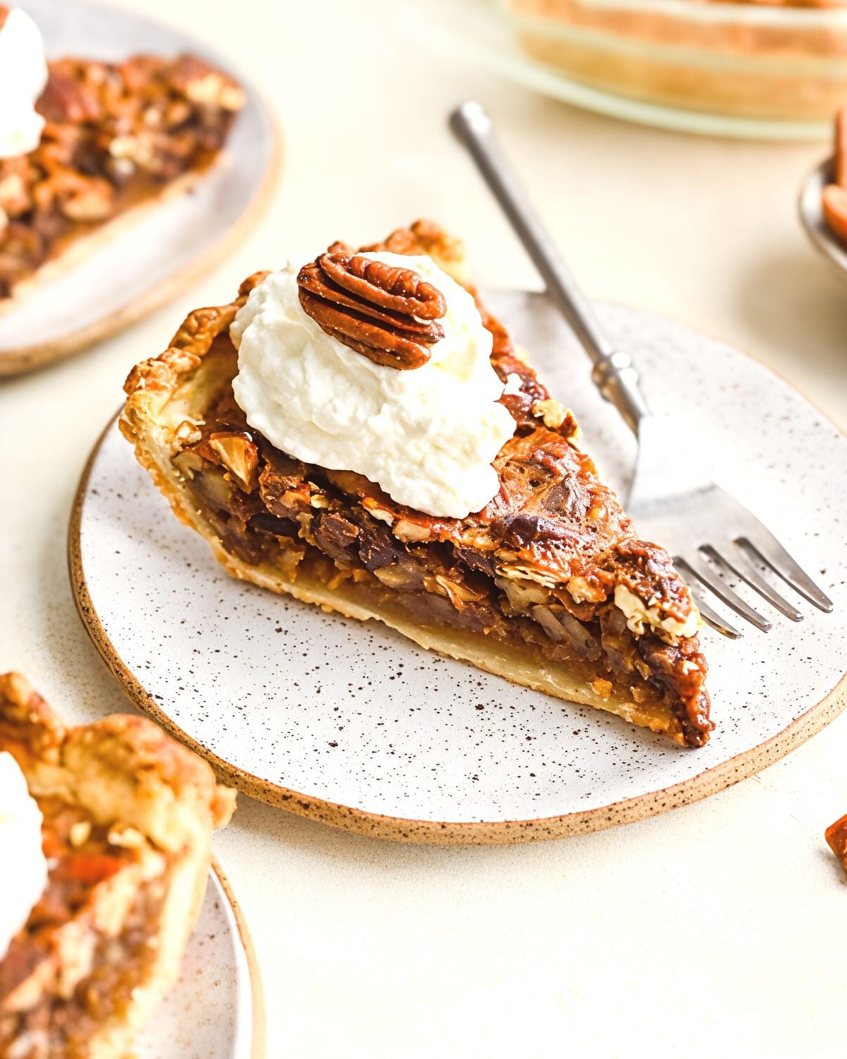 A slice of chocolate pecan pie topped with a dollop of whipped cream and a whole pecan rest on a speckled ceramic plate next to a dessert fork.