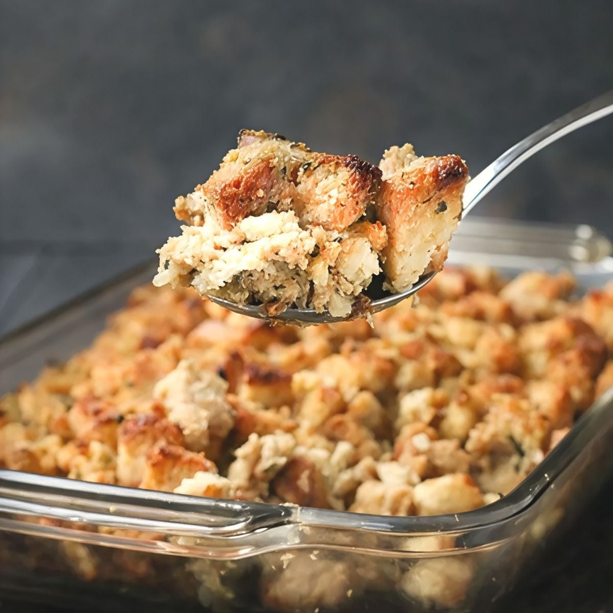 A metal serving spoon scooping a portion of golden, herb-seasoned bread stuffing from a glass baking dish