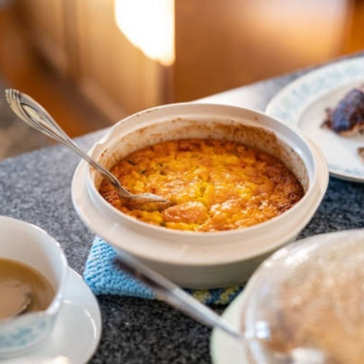 Corn Pudding’s Unsteady Center with a silver spoon inside and some additional dishes on the side