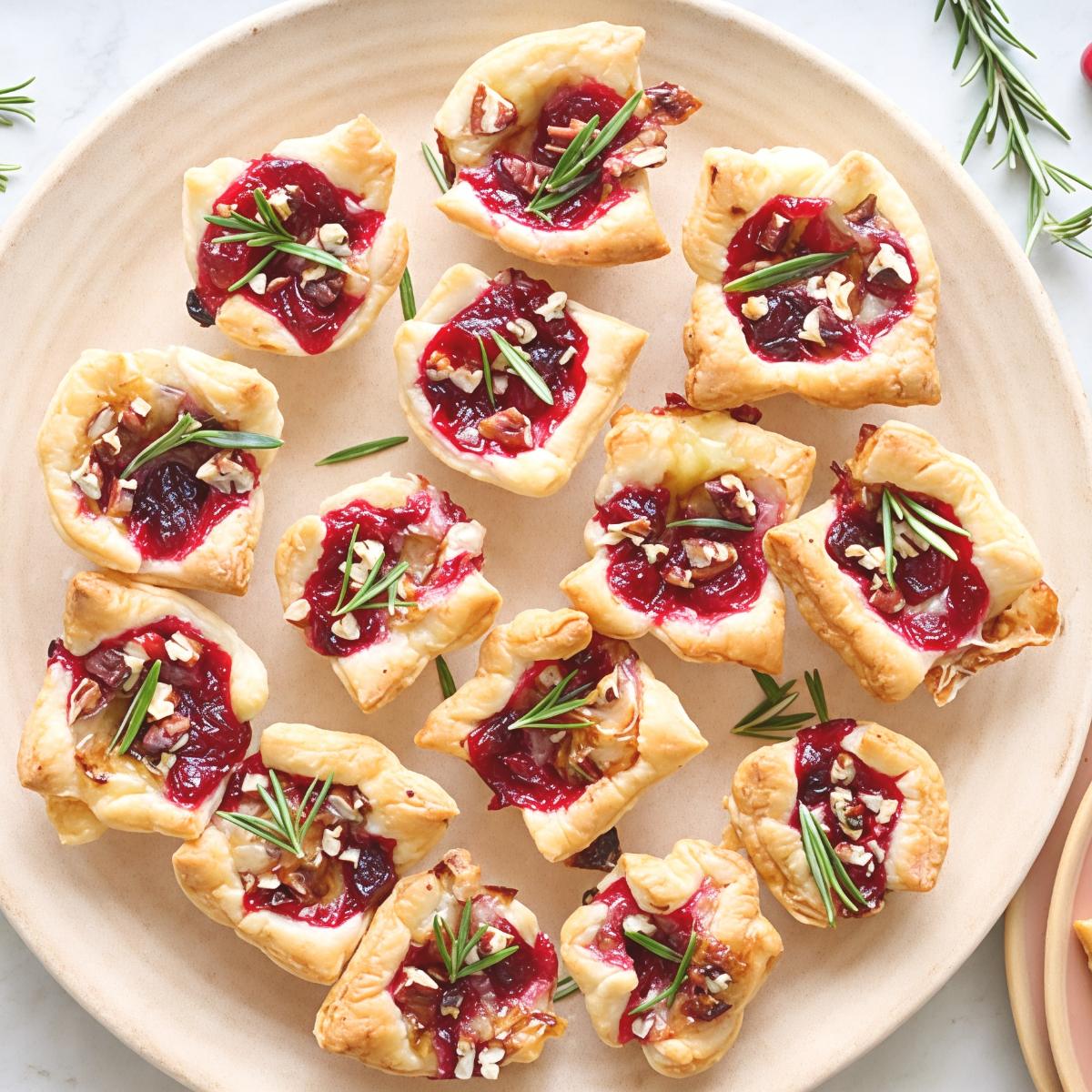 Golden puff pastry bites filled with melted Brie cheese and bright red cranberry sauce are arranged on a circular platter, garnished with chopped pecans and fresh rosemary sprigs.