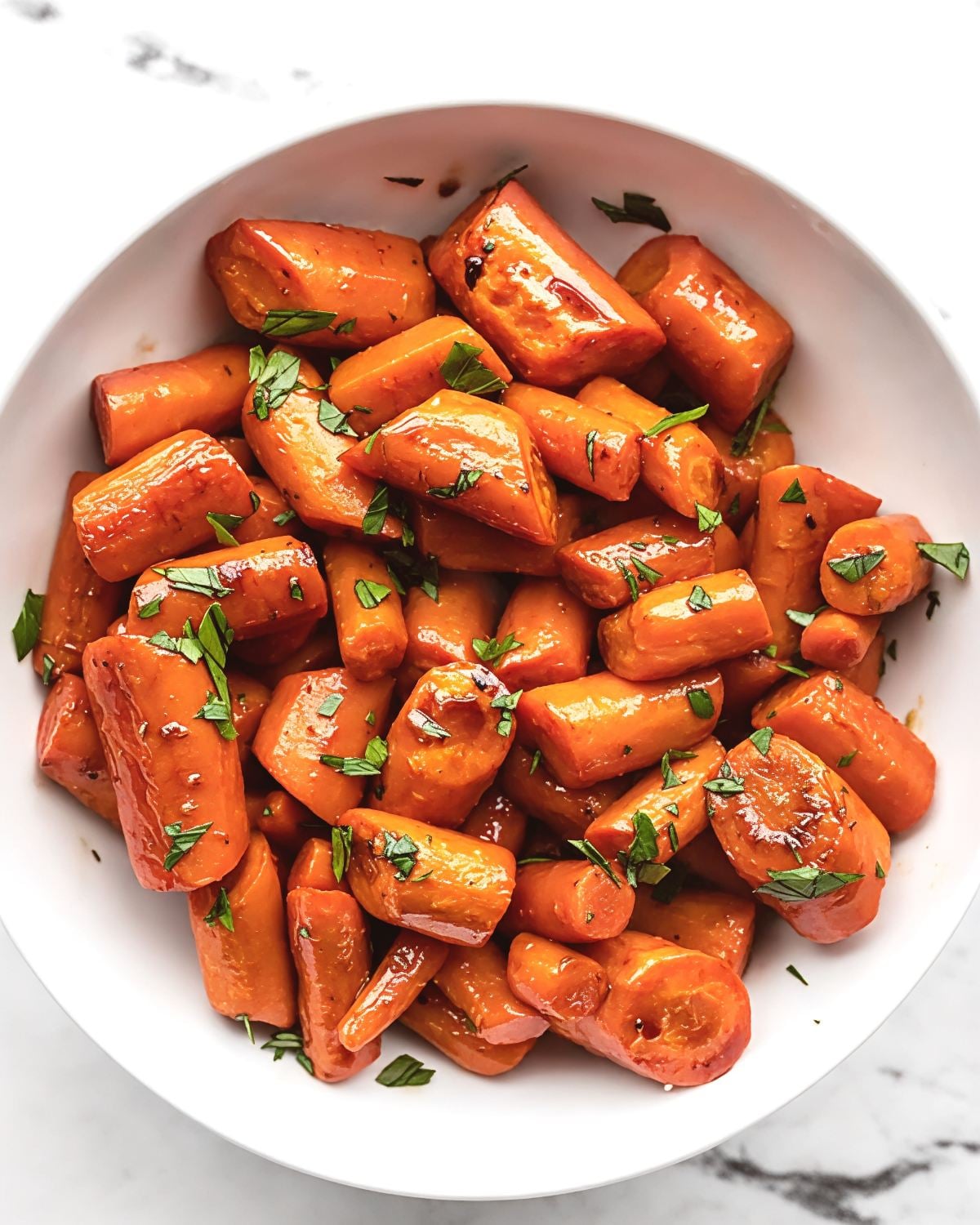 Tender roasted carrot chunks coated in a glossy honey balsamic glaze and sprinkled with chopped fresh parsley are piled in a white serving bowl.