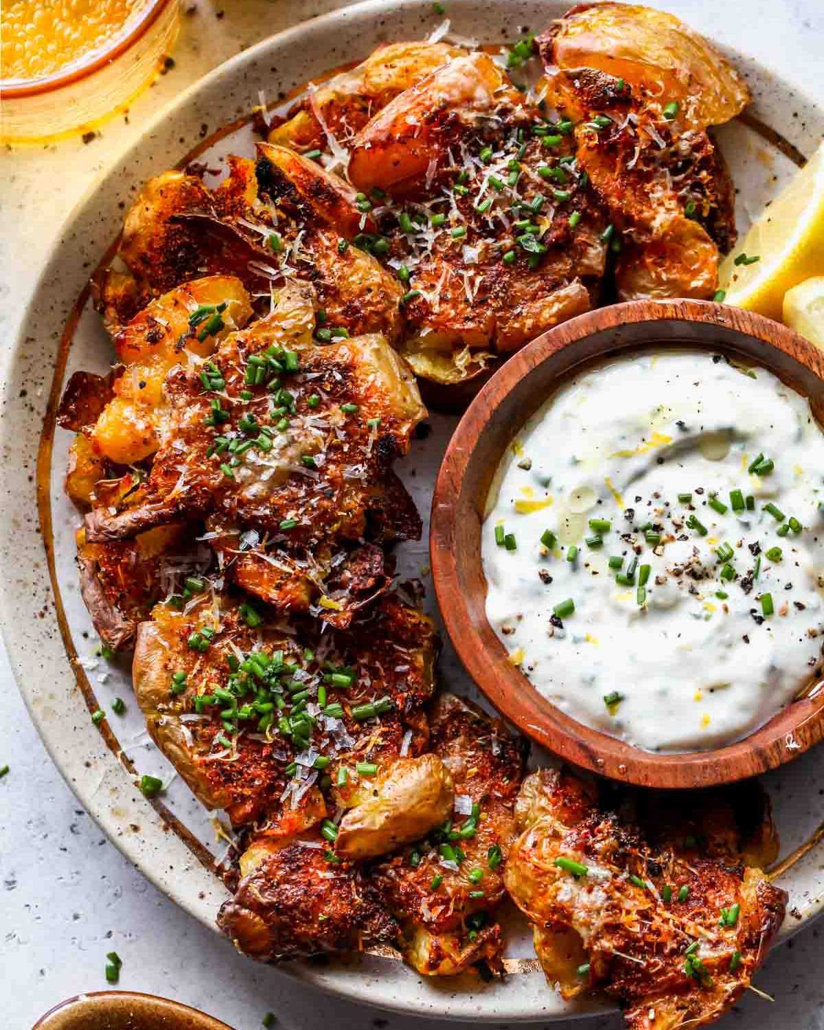 Crispy, paprika and Parmesan-seasoned smashed potatoes sprinkled with fresh chives are arranged on a platter alongside a wooden bowl of garlic aioli dip.