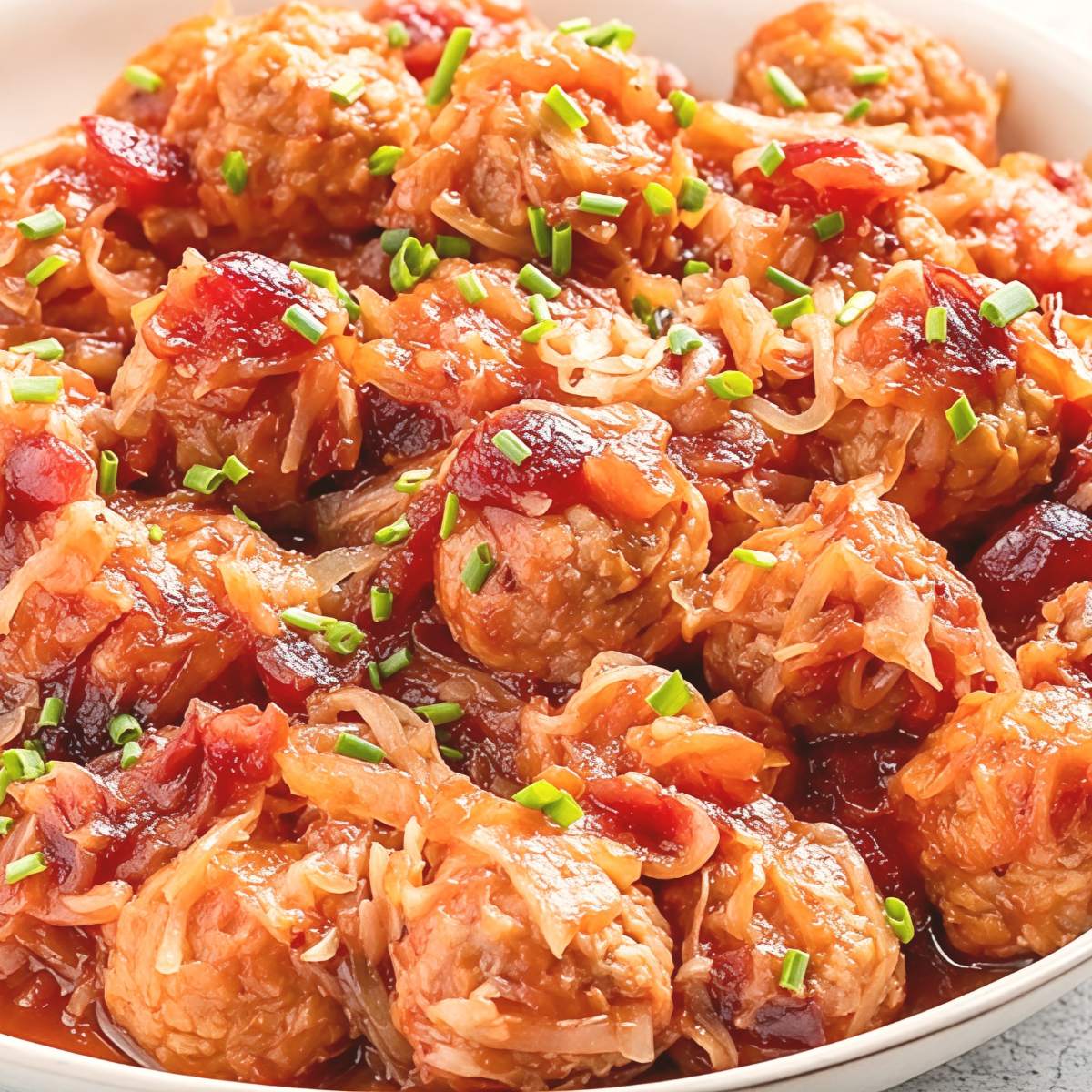 A bowl filled with cooked meatballs coated in a glossy cranberry sauce and sauerkraut, garnished with chopped fresh chives.