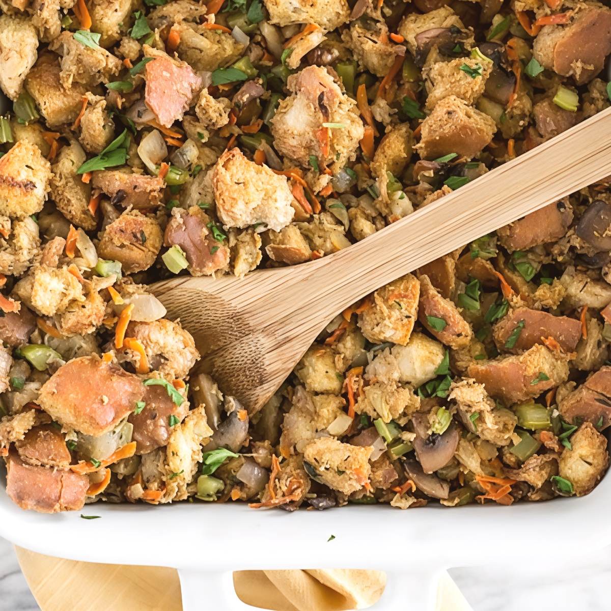 A white baking dish filled with golden baked vegetarian stuffing made with toasted bread cubes, herbs, diced carrots, celery, and mushrooms, complete with a wooden serving spoon.