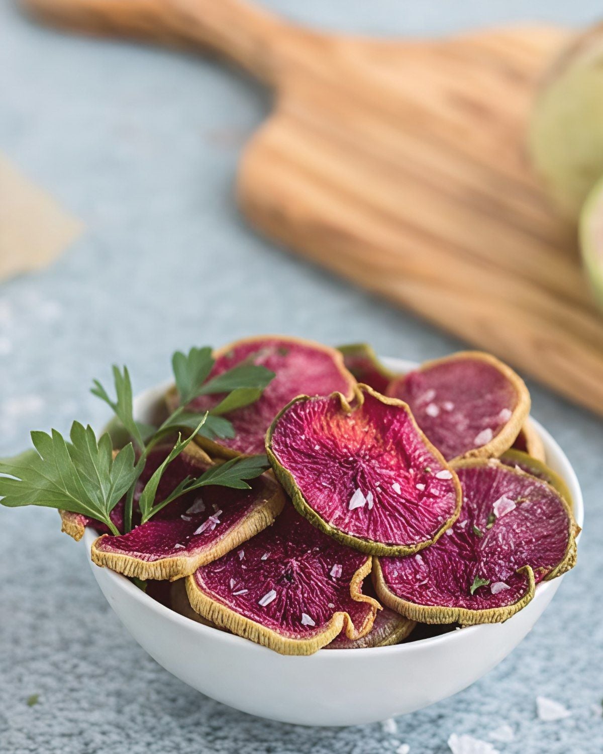 Crispy baked watermelon radish chips sprinkled with flaky sea salt and garnished with fresh parsley served in a white bowl for a healthy snack.