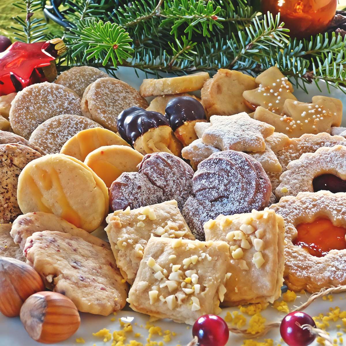 A close-up shot of a holiday platter filled with a variety of traditional Swiss Christmas cookies, including powdered star-shaped biscuits, jam-filled Linzer cookies, chocolate-dipped treats, and nut-topped squares, garnished with pine branches, hazelnuts, and red berries.