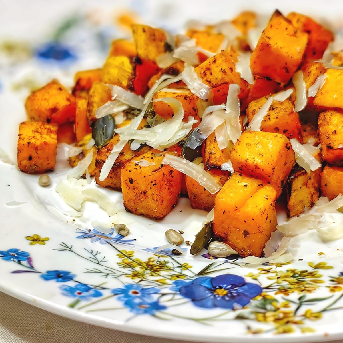 Seasoned air fryer roasted pumpkin cubes topped with shaved Parmesan cheese and pumpkin seeds on a floral plate for Thanksgiving vegetable side dishes.