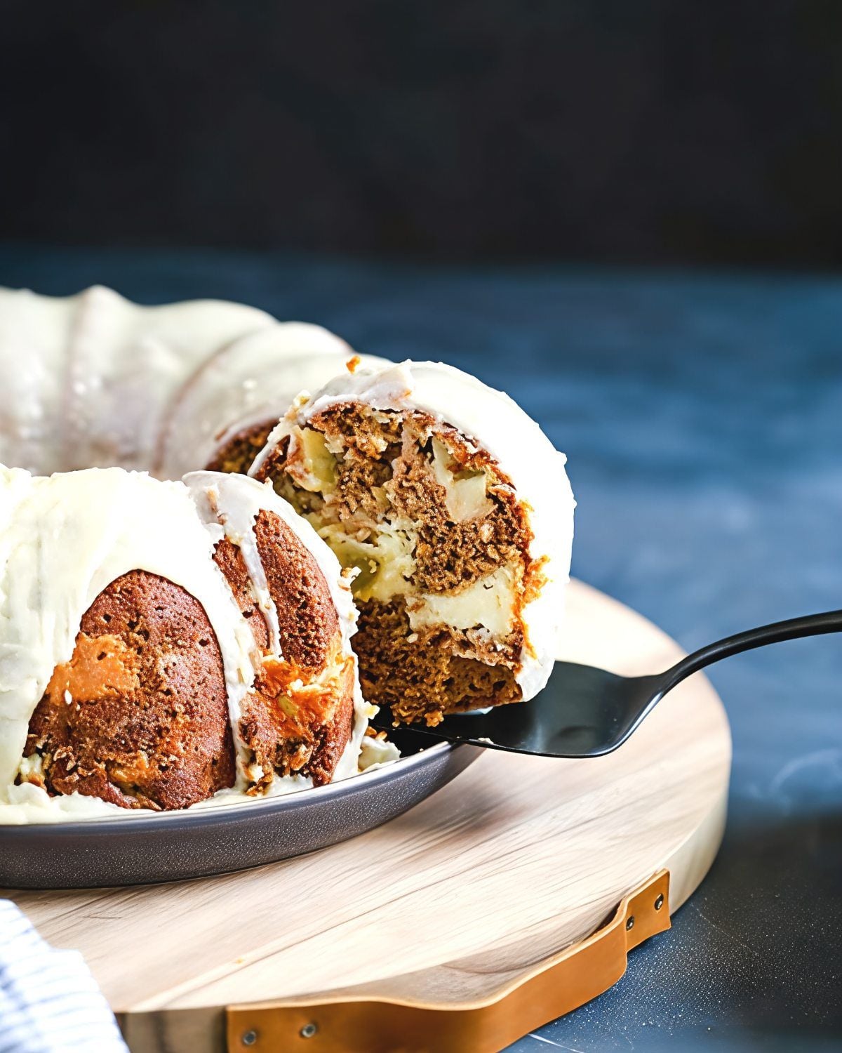 A cake server lifts a slice from a spiced apple Bundt cake topped with white cream cheese icing, revealing a cream cheese filling swirl inside.