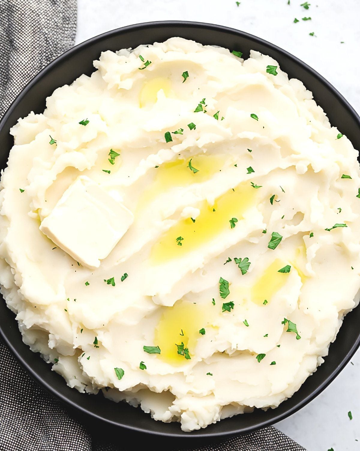 Best creamy vegan mashed potatoes in a dark bowl topped with vegan butter, olive oil, and fresh parsley for Thanksgiving.