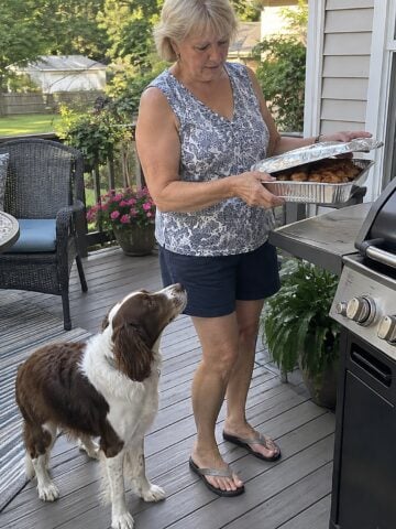 On a back porch with a grill in late afternoon, a woman lifts a foil pan from a side shelf while a dog stands underfoot, both caught mid-moment.