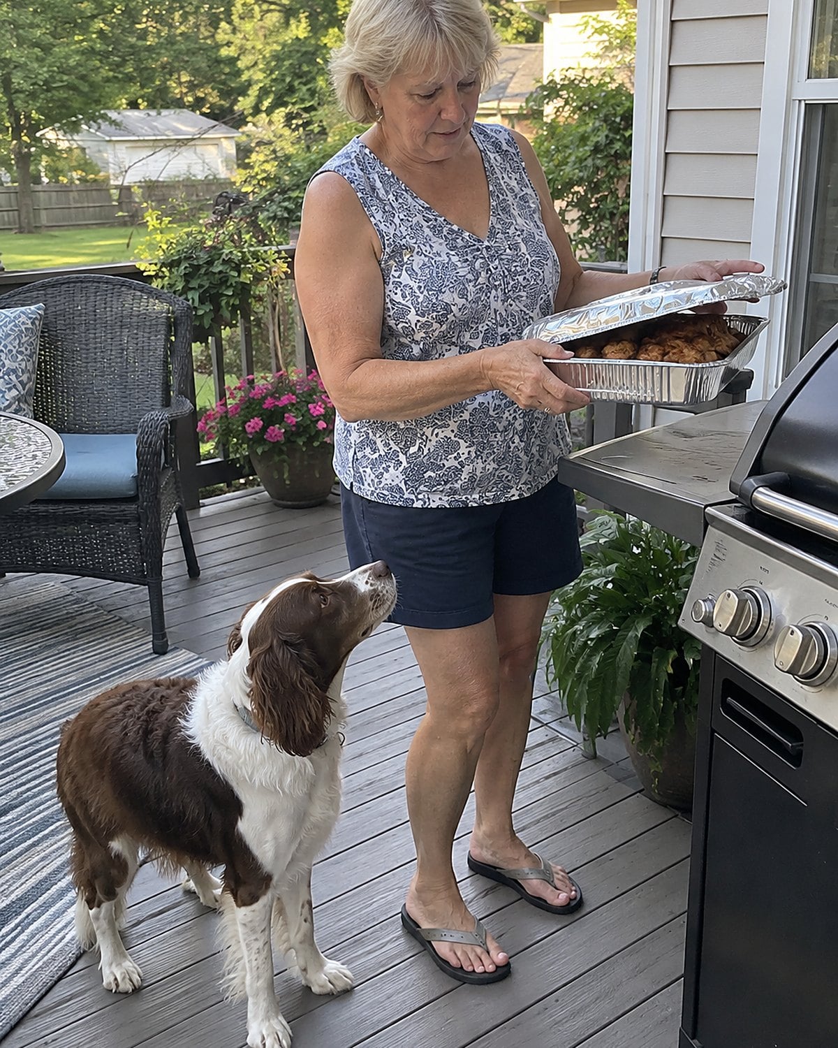 On a back porch with a grill in late afternoon, a woman lifts a foil pan from a side shelf while a dog stands underfoot, both caught mid-moment.