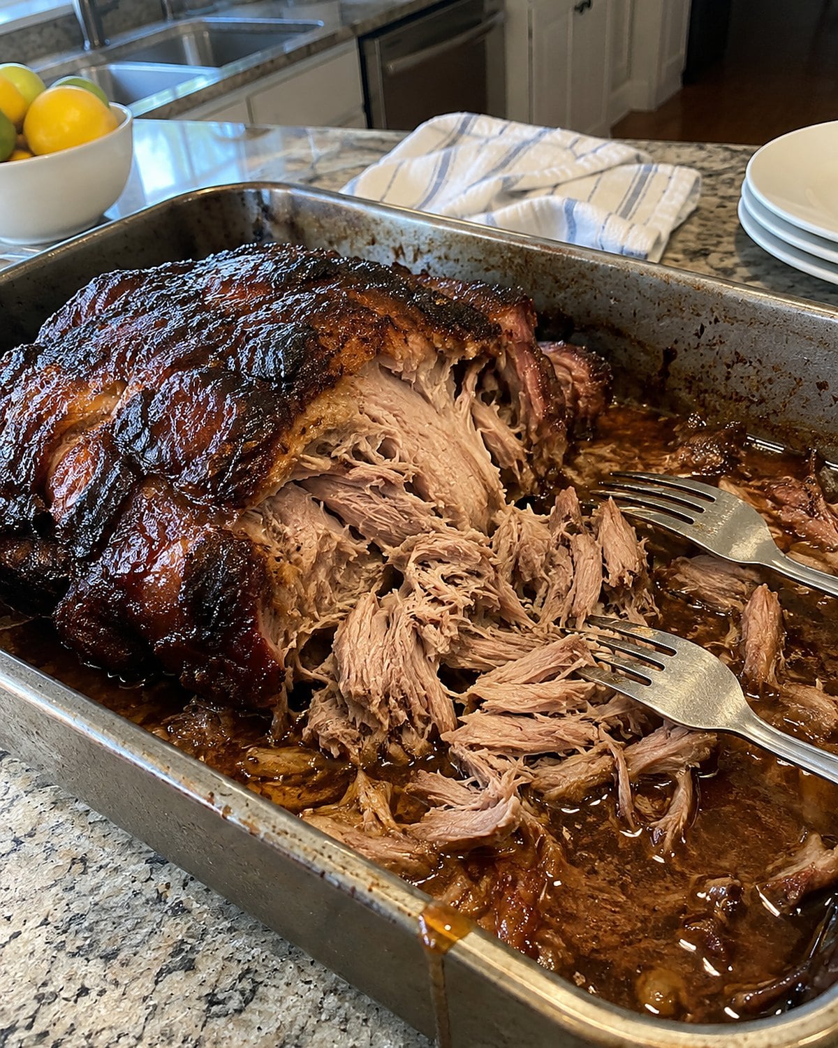 A partly pulled pork shoulder sits in its juices in a roasting pan, with forks beside it.