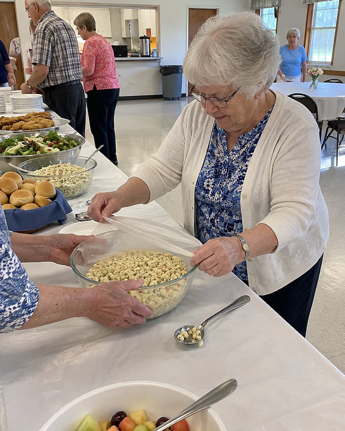 Hands cover a bowl of macaroni salad at the end of a church hall serving table.