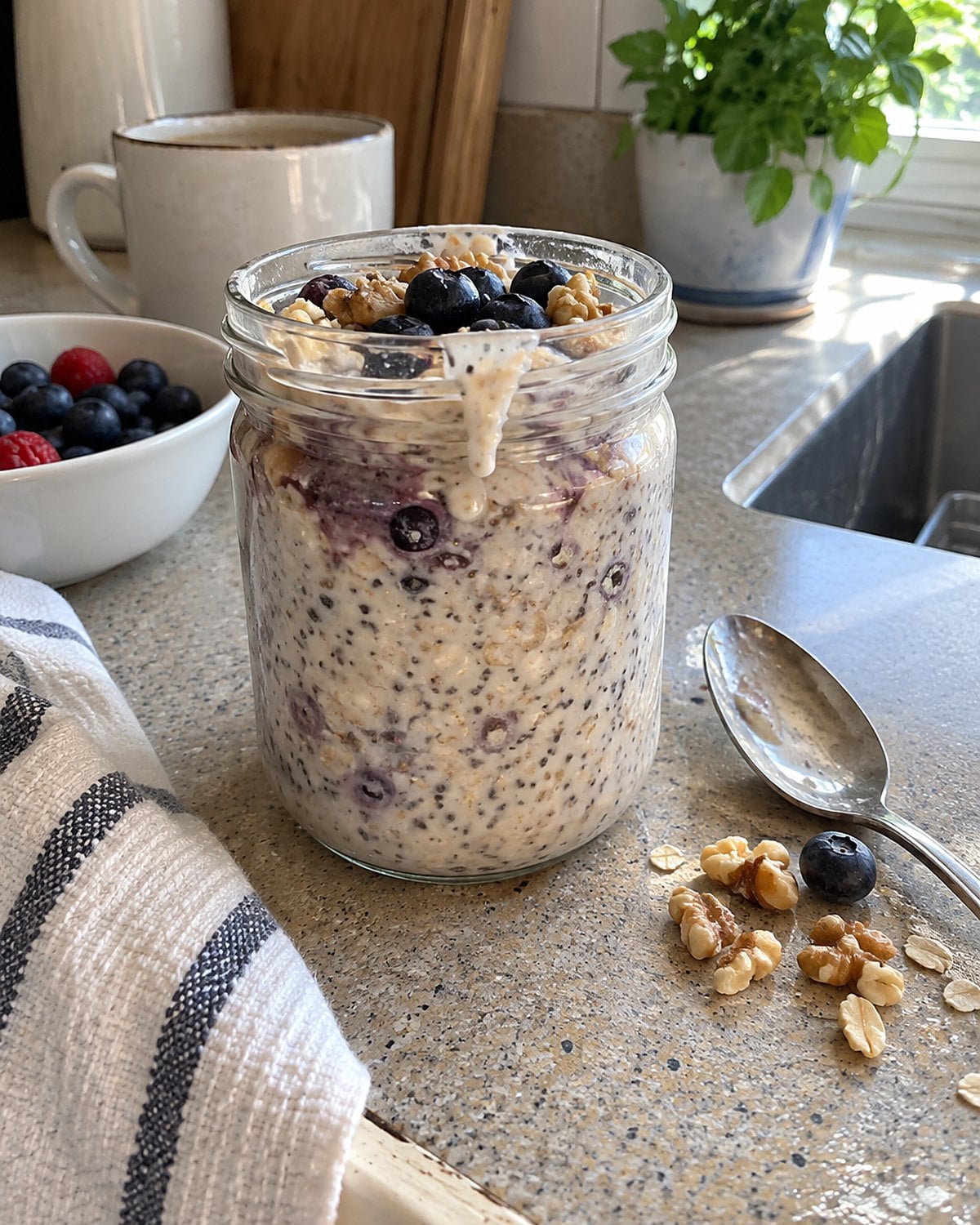 A small jar of overnight oats sits on a breakfast counter with a spoon and a few scattered nuts.