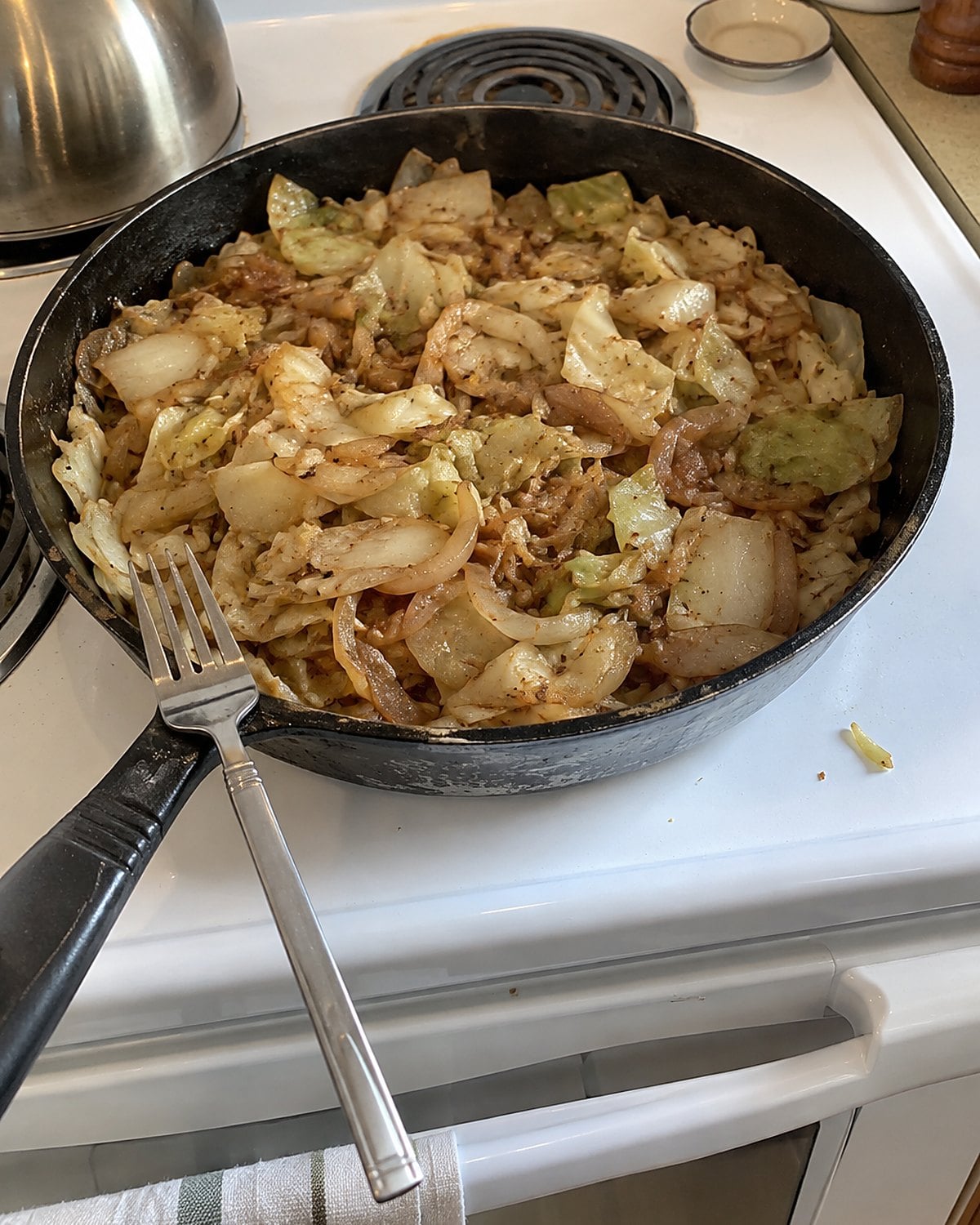 Braised cabbage and onion sit in a wide skillet on a white stove, with a fork resting on the handle.
