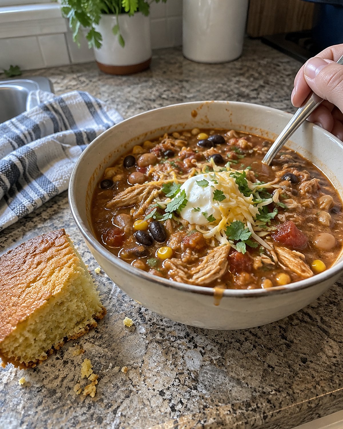 A bowl of chicken chili beside a rough-cut piece of cornbread on a kitchen counter, with a few crumbs between them.