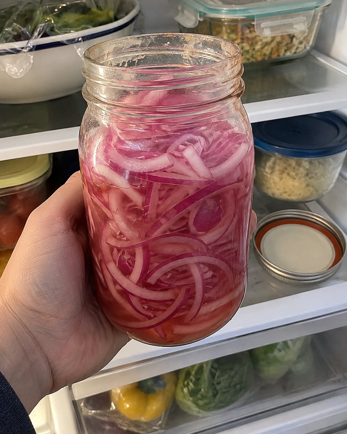 A jar of red onion slices sits on a fridge shelf beside covered leftover containers.