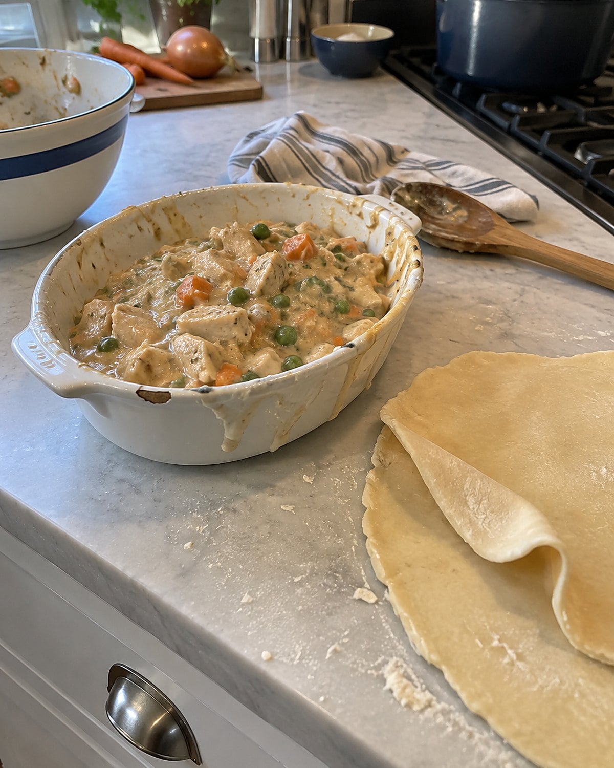 A small baking dish of chicken pot pie filling sits beside an unbaked pastry top on the counter.