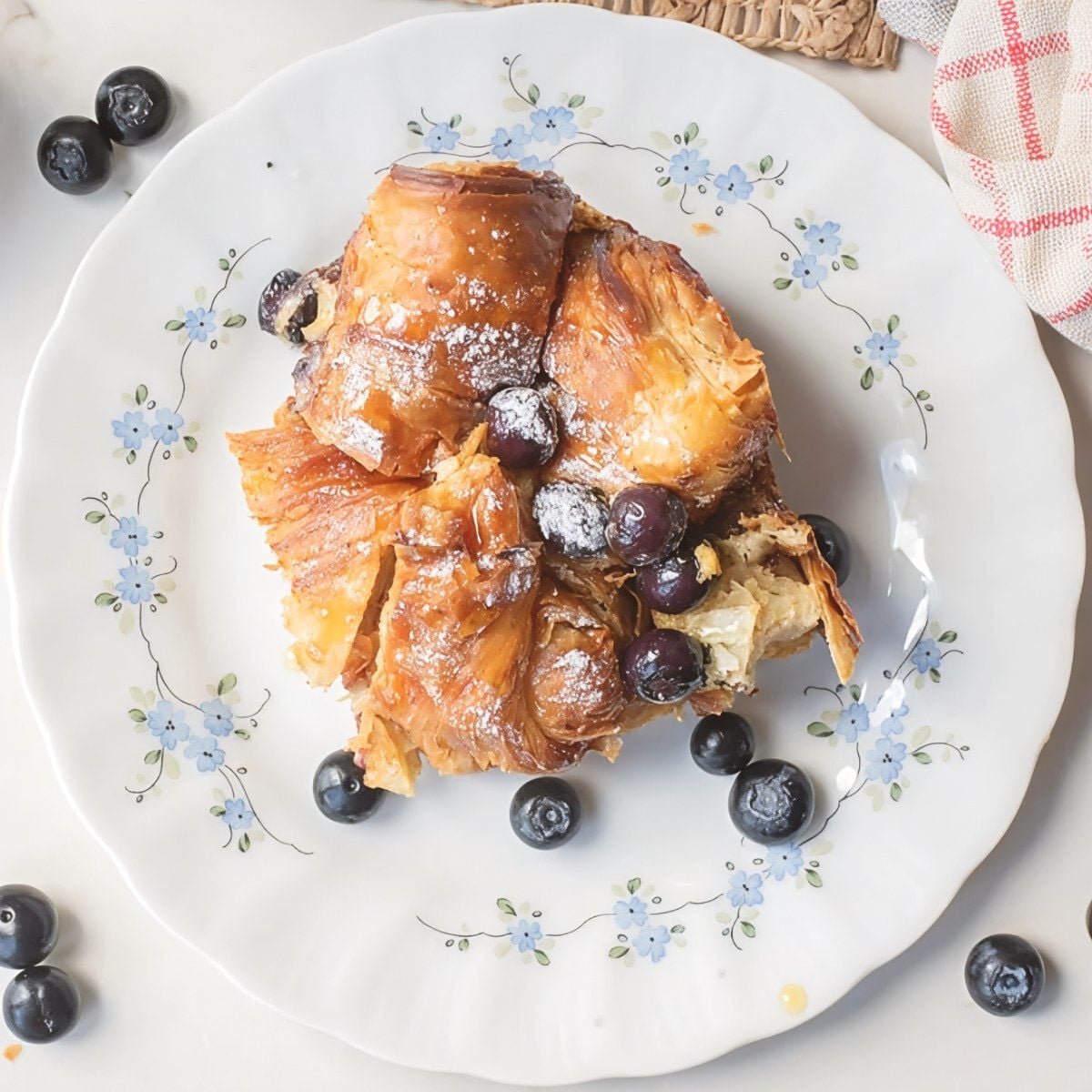 A slice of golden blueberry croissant french toast bake dusted with powdered sugar and topped with fresh blueberries served on a floral plate as Thanksgiving breakfast ideas.