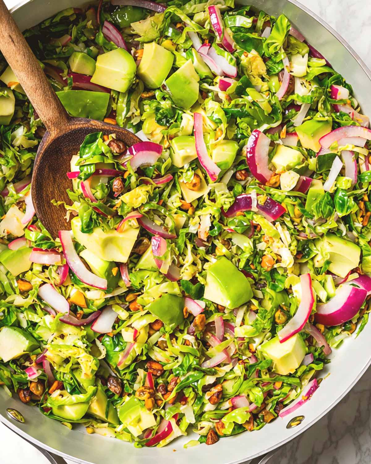 A large bowl filled with a fresh shaved Brussels sprouts salad tossed with diced avocado, thinly sliced red onion, and toasted nuts, served with a wooden spoon.