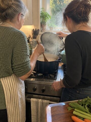 At a stove in late afternoon, a woman and her adult daughter lift a pot lid propped on a spoon and look into the simmering pot, neither facing the camera.