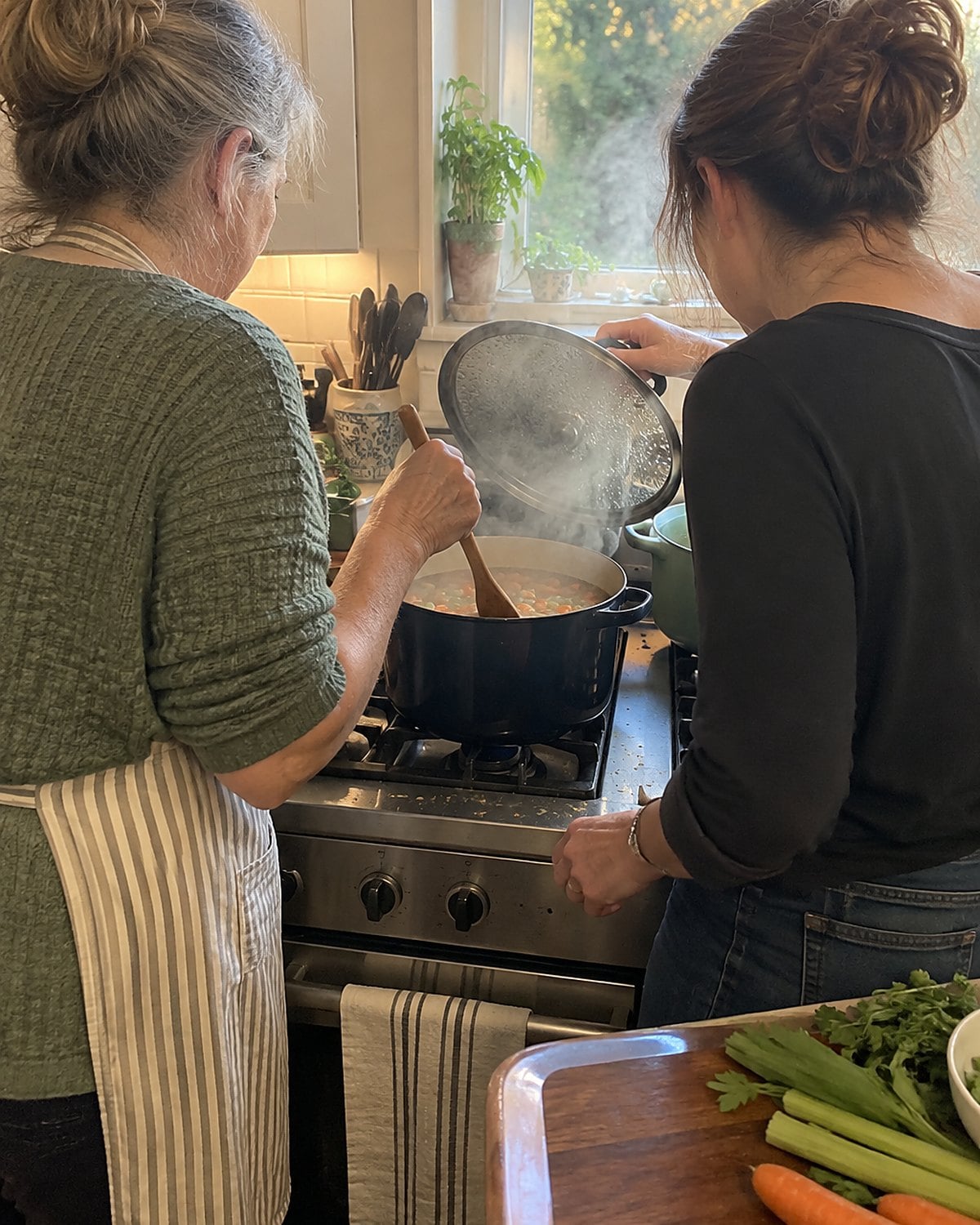 At a stove in late afternoon, a woman and her adult daughter lift a pot lid propped on a spoon and look into the simmering pot, neither facing the camera.