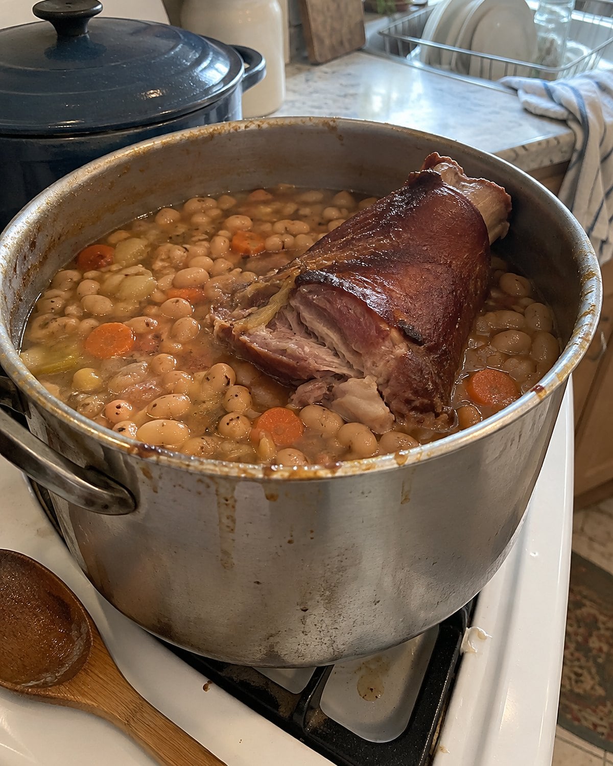 A smoked ham hock resting against the rim of a dented bean pot on a white stove.