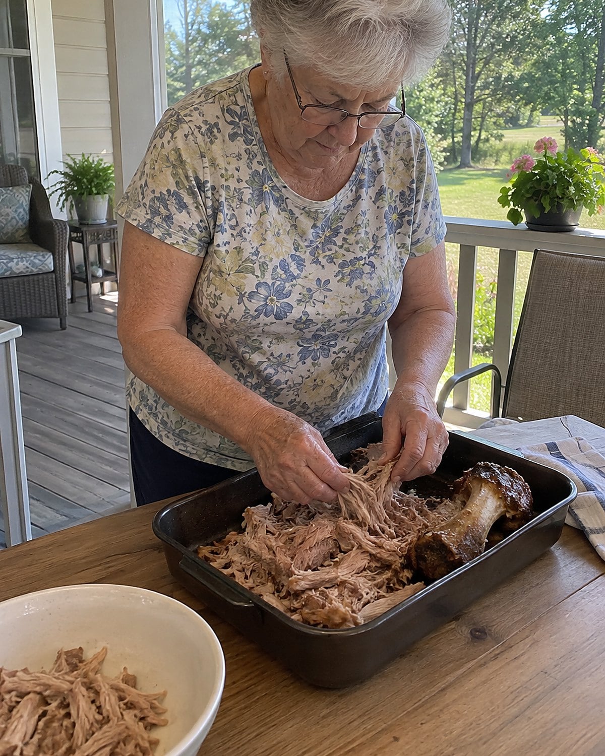 A hand pulling pork shoulder apart in a pan on a porch table.