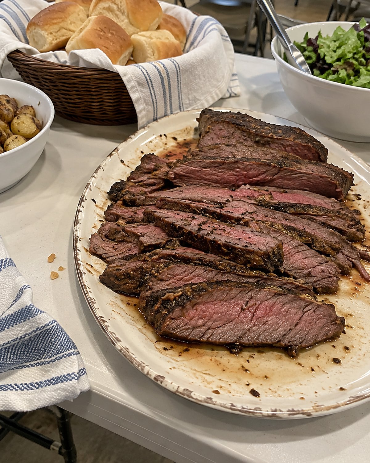 A slightly uneven platter of thin flank steak slices beside a basket of rolls on a church hall table.