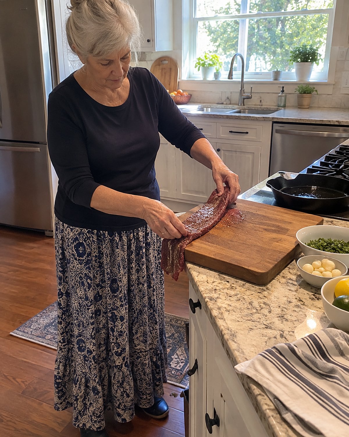 An older woman's hands turning a long skirt steak on a cutting board beside a cast-iron pan.