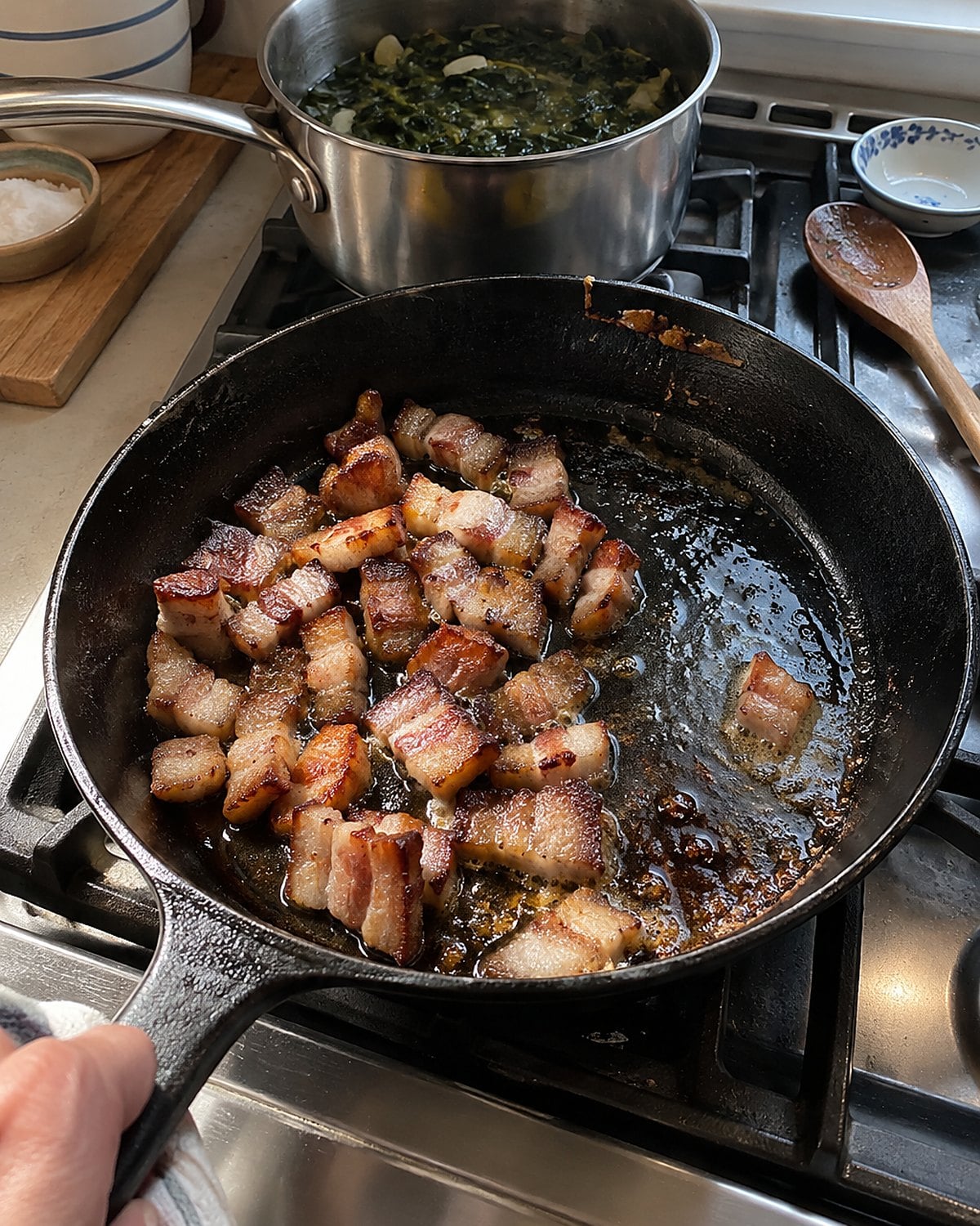 Small pieces of pork jowl browning in a skillet beside a pot of greens.