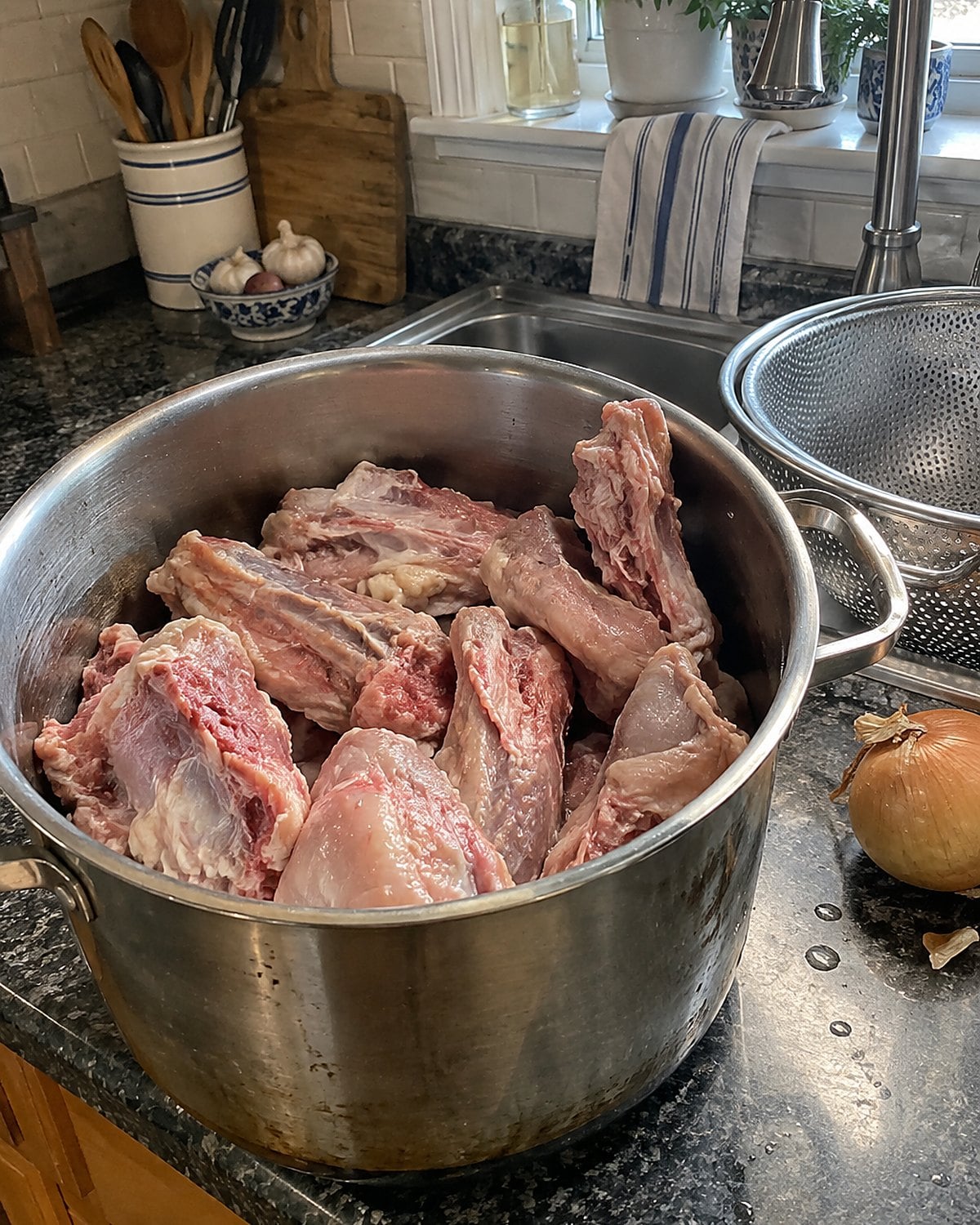 Chicken backs in a stockpot beside an onion and a wire strainer at the sink.
